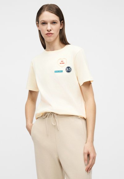 Γυναίκα που φοράει ανοιχτό κίτρινο T-shirt με τομπελάκι καρδιά "BE KIND", τομπελάκια "WEEKEND" και "83", και μπεζ παντελόνι με κορδονάκι, στέκεται με το χέρι στην τσέπη.