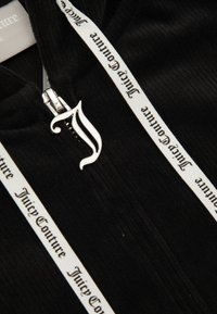 Casaquinho preto com fecho éclair, com textura canelada, apresentando um puxador de fecho prateado e cordão branco com o texto "Juicy Couture" em preto.