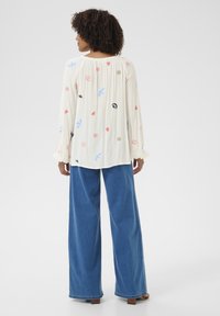 White long-sleeve blouse with colorful embroidered designs like hearts and birds, paired with loose blue wide-leg jeans.