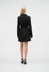 Γυναίκα με μακριά ξανθά μαλλιά, ντυμένη με μαύρο σακάκι-φόρεμα με ζώνη και μαύρες σανδάλες με τακούνι, γυρισμένη πλάτη σε λευκό φόντο.