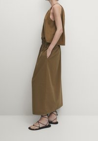 Haut brun sans manches, à coupe ample, associé à une longue jupe assortie avec une ceinture élastique et des poches latérales. Sandales noires à lanières.