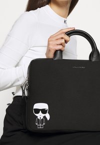 Sort håndtaske med glat stof, læderhåndtag, lynlås lukning og klistermærke af tegneseriefigur i hvid og sort på fronten.