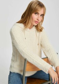 Maglione con cappuccio color crema realizzato in un morbido materiale peloso con un motivo texture. Presenta chiusura con cordini e maniche lunghe.