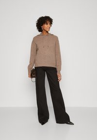 Felpa a maglia marrone con cappuccio e coulisse, abbinata a pantaloni neri a gamba larga. Gli accessori includono una piccola borsa nera. Texture liscia, design casual.