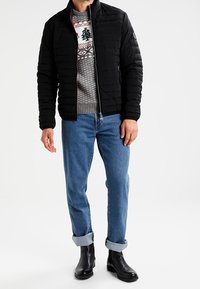 Svart puffjacka med hög krage bärs över en grå mönstrad tröja, kombinerat med ljusblå rullade jeans och svarta stövlar.