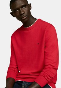 Maglione rosso a collo tondo realizzato in tessuto knit, con polsini e orlo a coste, dotato di un piccolo logo sul petto. Indossato con pantaloni in denim scuri.