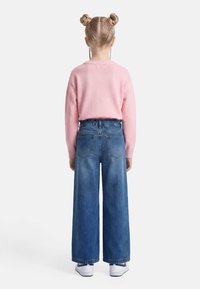 Παιδί με διπλό κότσο στα μαλλιά, ντυμένο με ροζ πουλόβερ, φαρδιά μπλε τζιν και λευκά αθλητικά, στέκεται με την πλάτη προς τα εμπρός.