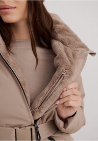 Beige jakke med pelsforet krave, der har lynlås og b�ælte i taljen. Den glatte yderflade komplementerer den bløde indre tekstur.