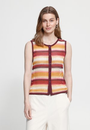 Donna con capelli castano chiaro indossa un gilet lavorato a uncinetto senza maniche a righe multicolori e pantaloni bianchi, in piedi davanti a uno sfondo grigio chiaro.