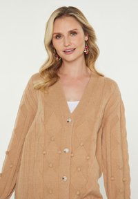 Blonde Frau, die einen beigen Pullover mit Zopfmuster und Knöpfen trägt, sowie farbenfrohen, hängenden Ohrringen, lächelt vor einem schlichten hellen Hintergrund.