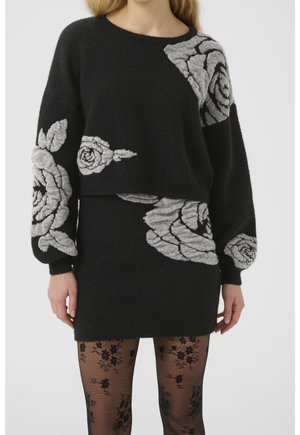 Femme portant un ensemble pull et jupe en tricot noir avec de grands motifs de roses grises, associée à des collants noirs transparents à motifs floraux.