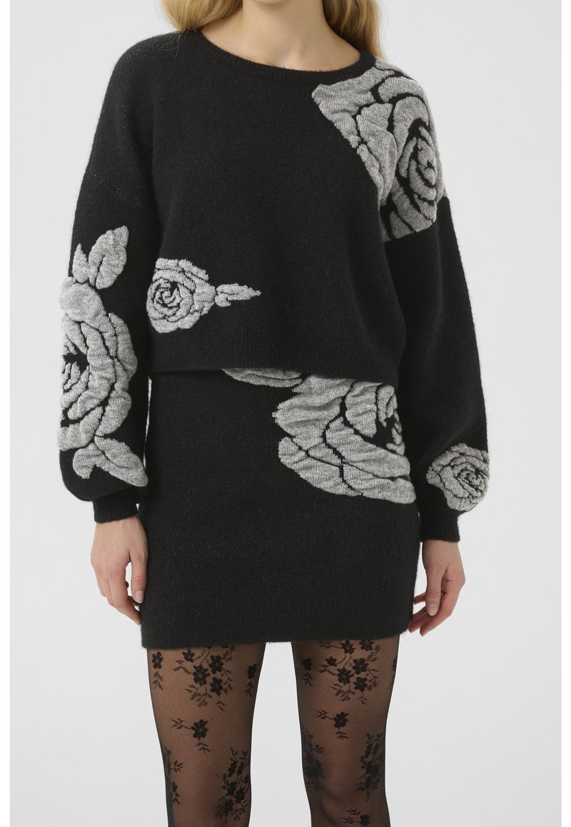 Femme portant un ensemble pull et jupe en tricot noir avec de grands motifs de roses grises, associée à des collants noirs transparents à motifs floraux.