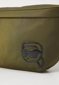 Olivenfarbene Nylon-Tasche mit einem glänzenden schwarzen Logo eines Gesichts, das eine Sonnenbrille und einen Pferdeschwanz trägt, mit einer glatten, eleganten Textur.