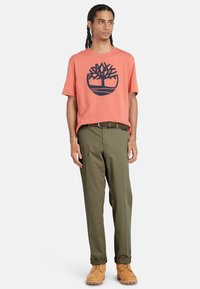 Coral T-shirt with a navy tree logo, paired with olive green pants and tan boots. The outfit features a relaxed fit and cotton fabric.