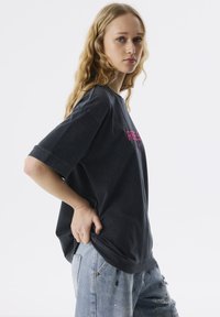 Black oversized t-shirt with short sleeves, featuring pink text. Worn with faded blue distressed jeans, showcasing a relaxed fit and casual style.