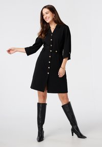 Zwart shirtjurk met driekwart mouwen, gouden knopen en een ceintuur in de taille. Draagt met zwarte leren laarzen tot de knie. Glad oppervlak.