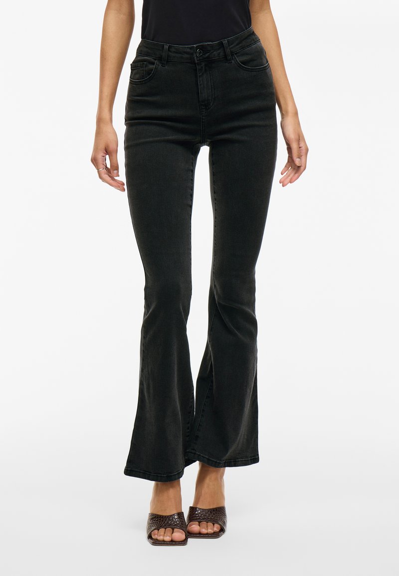 Jeans acampanados negros de denim, con cintura alta, cinco bolsillos y una textura suave. Combinados con sandalias de tacón abierto marrones.