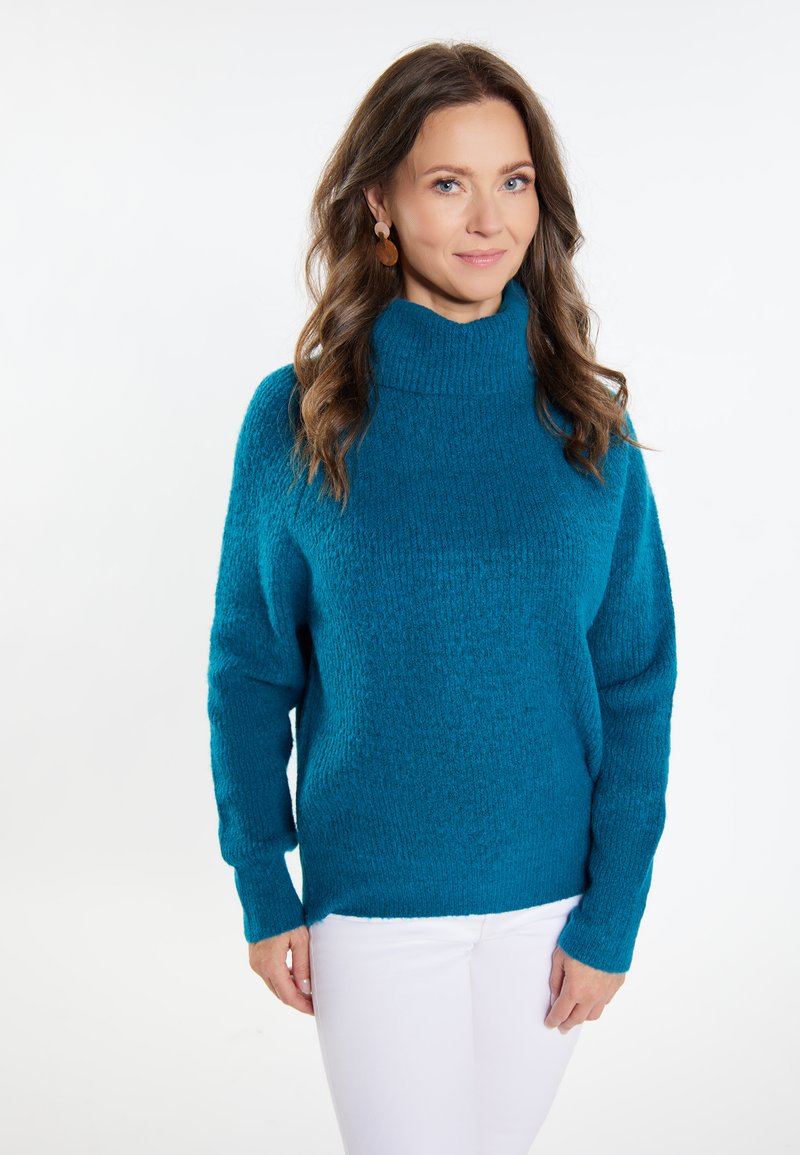 Teal, gestructureerde gebreide trui met een hoge, geribbelde halslijn en lange mouwen. Gecombineerd met witte broeken met een relaxte pasvorm en zijsplitten.