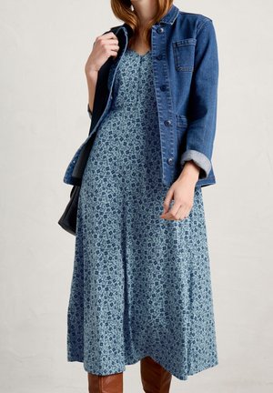 Femme portant une robe midi fleurie bleue avec une veste en denim, des bottes marron, et un sac noir en bandoulière.