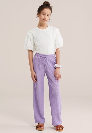 Młoda kobieta stojąca, ubrana w biały top z bufkami na rękawach, lawendowe, szerokie spodnie o fakturze, biały scrunchie na nadgarstku i brązowe sandały.