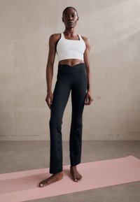 Fekete, magas derekú leggings és fehér sportmelltartó. A leggings sima textúrájú és enyhén bootcut szabású. A háttér neutrális és világos.