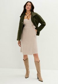 Giacca verde oliva, vestito avvolgente beige in maglia con cintura in vita, stivali marroni alla coscia e una sciarpa gialla a fantasia attorno al collo.