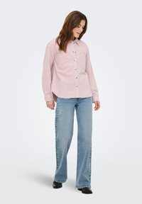 Jonge vrouw met bruin haar draagt een lichtroze overhemd, blauwe wijde jeans en zwarte schoenen, staand tegen een witte achtergrond.