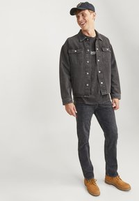 Chaqueta de mezclilla negra con bolsillos frontales, combinada con una camiseta gris de estampado, jeans negros y botines marrones con cordones. Diseño de atuendo casual.