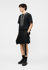 Νεαρός άνδρας με σγουρά μαλλιά που φοράει μαύρο κοντομάνικο πουκάμισο με διακοσμητικά κορδόνια, μαύρα σορτς με υφασμάτινα κορδόνια, μαύρες κάλτσες και μαύρα αθλητικά παπούτσια.