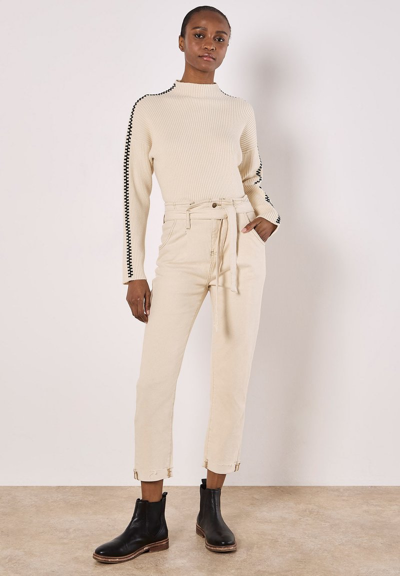 Cremfarbener gerippter Pullover mit schwarzen Seitendetails, kombiniert mit hochtaillierten, hellbeigen, tapered Pants. Schwarze Stiefeletten runden das Outfit ab.