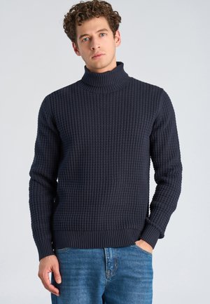 Young man with curly hair wearing a dark ribbed turtleneck sweater and blue jeans, standing with one hand in pocket against plain background.