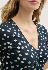 Dunkelblaues Kleid mit einem Muster aus hellblauen und weißen Punkten. V-Ausschnitt mit gerafften Details an der Brust. Weicher, dehnbarer Stoff.