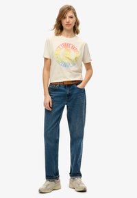 Camiseta de manga corta color crema con un gráfico circular multicolor, combinada con jeans de mezclilla azules. El modelo lleva zapatillas grises claras y un cinturón marrón.