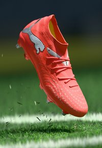 Φωτεινό πορτοκαλί ποδοσφαιρικό παπούτσι με υφασμένο επάνω μέρος, ασημί λογότυπο και διάφανα καρφιά, εμφανιζόμενο στον αέρα πάνω από πράσινο γρασίδι.