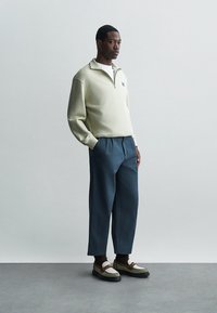 Šviesiai žalia, zip-up puloveris su balta apykakle, derinamas su tamsiai mėlynomis klostuotomis kelnėmis. Avalynė pasižymi įvairių tekstūrų ir spalvų deriniu.