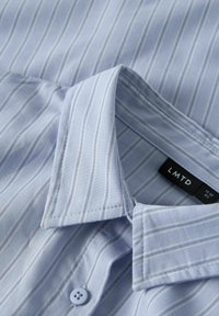 Camicia azzurra chiaro con strisce verticali bianche, colletto abbottonato e etichetta nera con la scritta "LMTD" che indica la taglia XS.