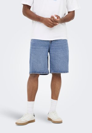 Persona con camiseta blanca, pantalones cortos vaqueros azules hasta la rodilla, calcetines blancos y zapatillas blancas, de pie frente a un fondo liso y claro.