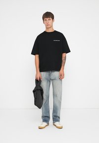Zwarte katoenen t-shirt met korte mouwen, met witte tekst "BESPAAR CULTUUR"; gecombineerd met lichte spijkerbroek en witte sneakers.