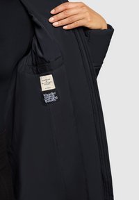 Sort dunjakke med en glat tekstur, der har et mærke og logo indeni. Jakken har et tætsiddende design med synlige syningsdetaljer.