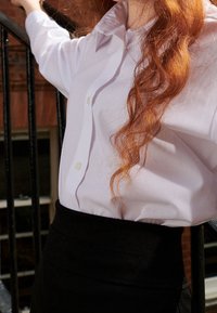Chemise lavande claire à boutons avec col pointu et manches retroussées, associée à une jupe noire cintrée, présentée contre une rambarde.