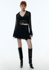 Zwarte mesh lang mouw cropped top met een v-hals en strikdetail aan de voorkant, gecombineerd met een zwarte rok met veters aan de zijkant en zwarte kniehoge laarzen.