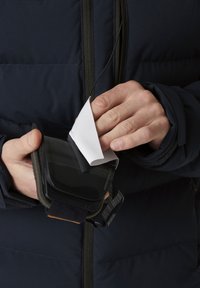 Schwarzes Touchscreen-Gerät, das in einer Hand gehalten wird, bedeckt mit einer flexiblen weißen Folie. Gerät mit einem dunklen Riemen gesichert, der sich kontrastierend zu einer marineblauen Jacke abhebt.