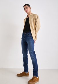 Camisa de manga larga negra sobre una camiseta sin mangas negra, combinada con unos jeans ajustados azul y zapatillas marrones. Texturas suaves, diseño casual.