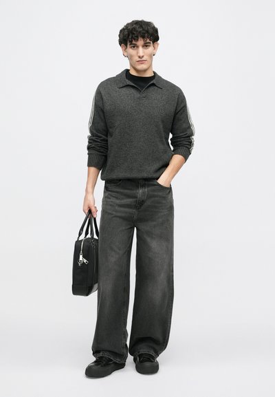 Grijze coltrui, losse zwarte jeans en zwarte schoenen. Houdt een zwarte tas vast met zilveren accenten. Eenvoudig en ruim ontwerp.