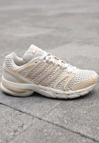 Weißer und beiger Sportschuh mit Mesh-Obermaterial und mehreren Streifen, auf grauem, gefliestem Boden platziert.