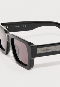 Czarne prostokątne okulary przeciwsłoneczne o matowym wykończeniu, z fioletowymi soczewkami i wzorzystym srebrnym akcentem na zausznikach.