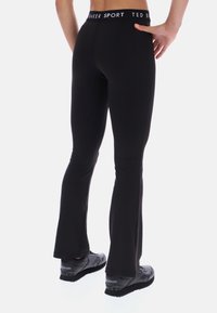 Čierne športové legíny so strečovým pásom, na ktorom je značka "TED BAKER SPORT". Priliehavý strih s rozšíreným lemom, v kombinácii s čiernymi športovými topánkami.