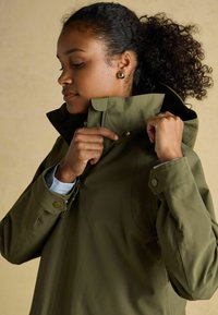 Jachetă impermeabilă de culoare verde măslin, cu un guler înalt, capse și material texturat. Dispune de un fermoar frontal și nasturi la mansete.