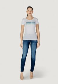 Fehér, testhezálló póló, amelyen csillogó türkiz színű "GUESS" felirat található, sötétkék skinny farmernadrággal és meztelen színű magassarkúval párosítva. Egyszerű dizájn, sima textúra.