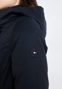 Mørkeblå jakke med hette, med et teksturert logopatch på venstre erme. Materialet ser holdbart ut med en glatt overflate.